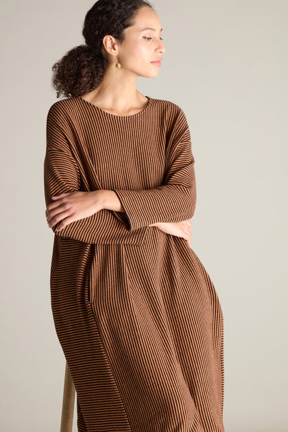 A person wearing a relaxed drop shoulder design, Mini Stripe Flared Dress in brown and black vertical stripes, sitting on a stool with arms crossed and looking to the side against a plain background.