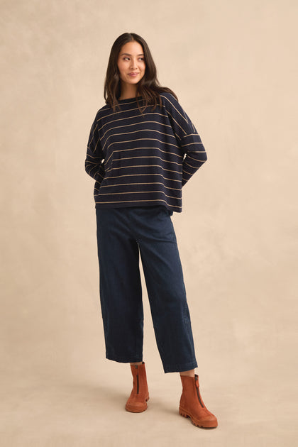 A woman wearing the Narrow Stripe Boxy Top in navy, styled with wide-leg dark blue jeans and brown ankle boots, stands against a beige background.