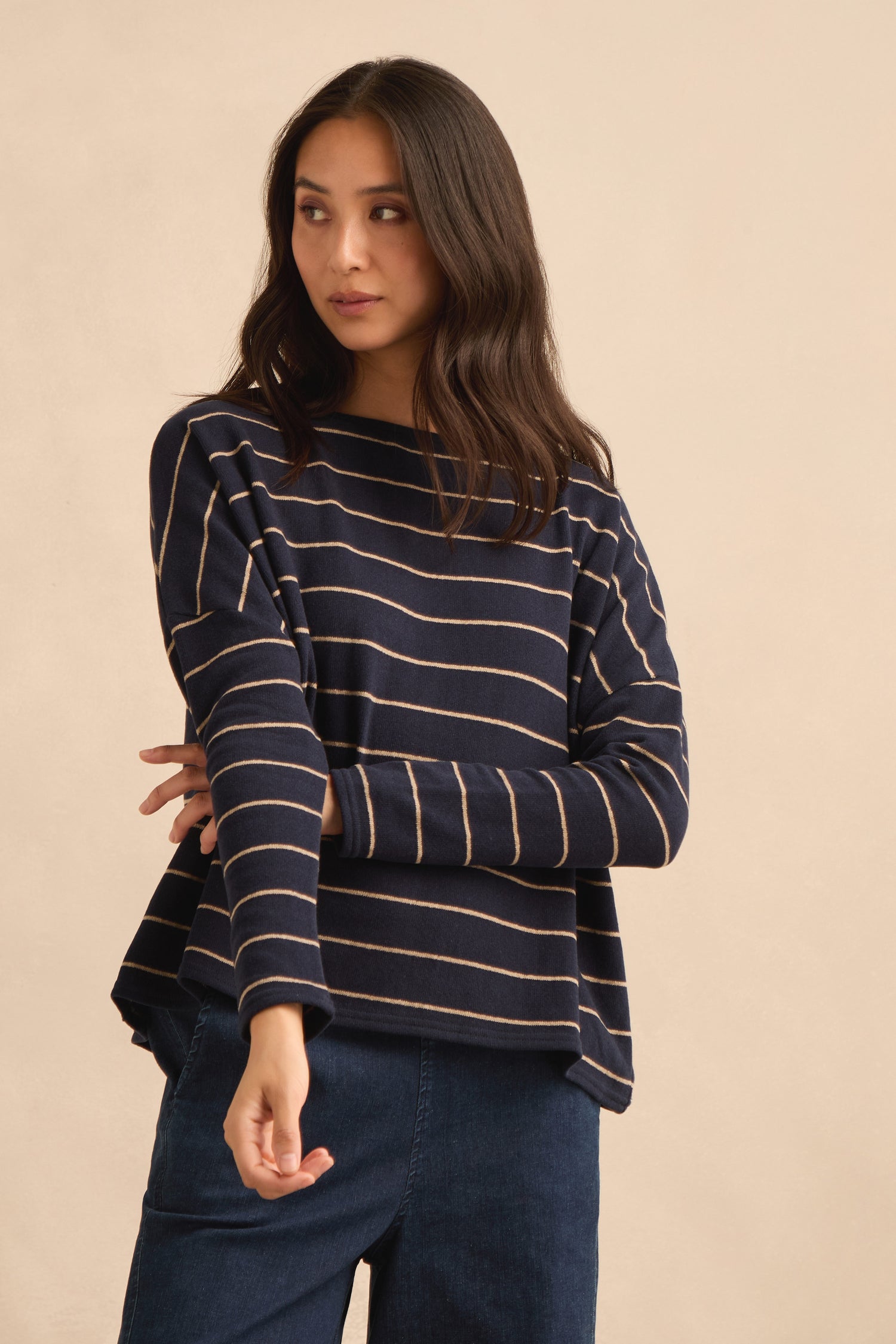 A woman stands against a plain beige background, wearing the Narrow Stripe Boxy Top—a navy blue, long-sleeve boxy fit top with beige horizontal stripes made in Italy—paired with blue pants.