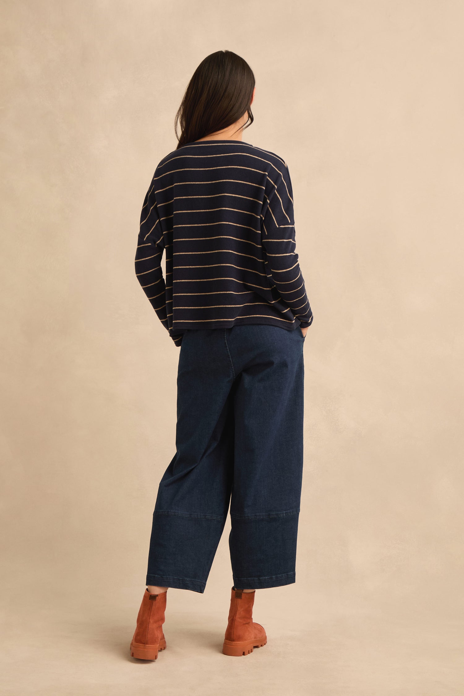 A person stands facing away, wearing the Narrow Stripe Boxy Top in navy stripes—featuring a boxy fit and crafted in Italy—paired with wide-leg dark blue jeans and brown ankle boots, set against a plain neutral background.