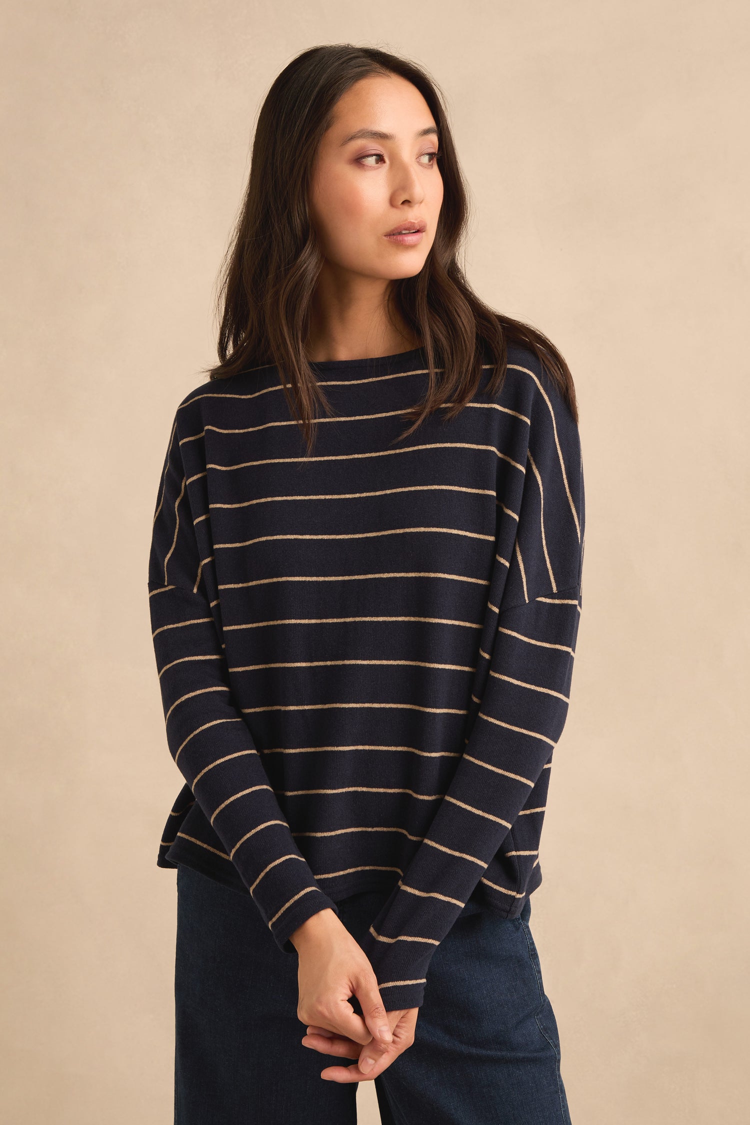 A woman wears the Narrow Stripe Boxy Top, a navy blue sweater with beige horizontal stripes and a relaxed, boxy fit, paired with blue pants. She stands against a beige background, gazing to the side—an effortlessly chic look made in Italy.
