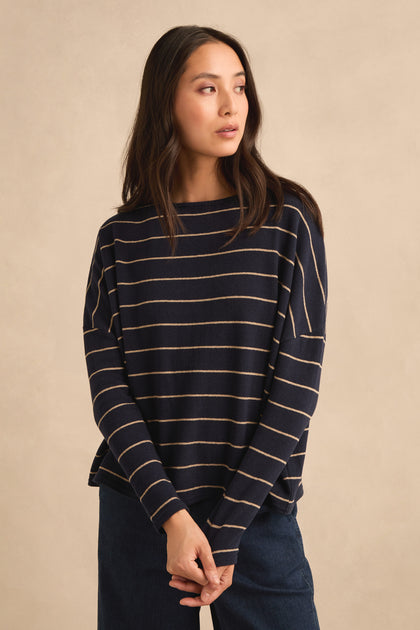 A woman wears the Narrow Stripe Boxy Top, a navy blue sweater with beige horizontal stripes and a relaxed, boxy fit, paired with blue pants. She stands against a beige background, gazing to the side—an effortlessly chic look made in Italy.