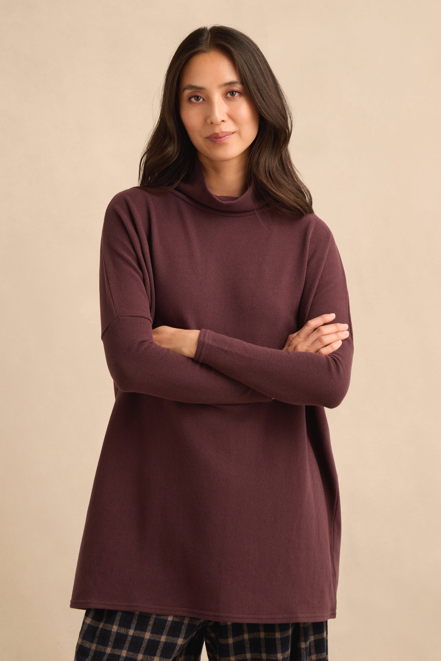 A woman with long dark hair stands with her arms crossed against a plain beige background, wearing a Cowl Neck Fig Fleece Tunic and plaid pants.