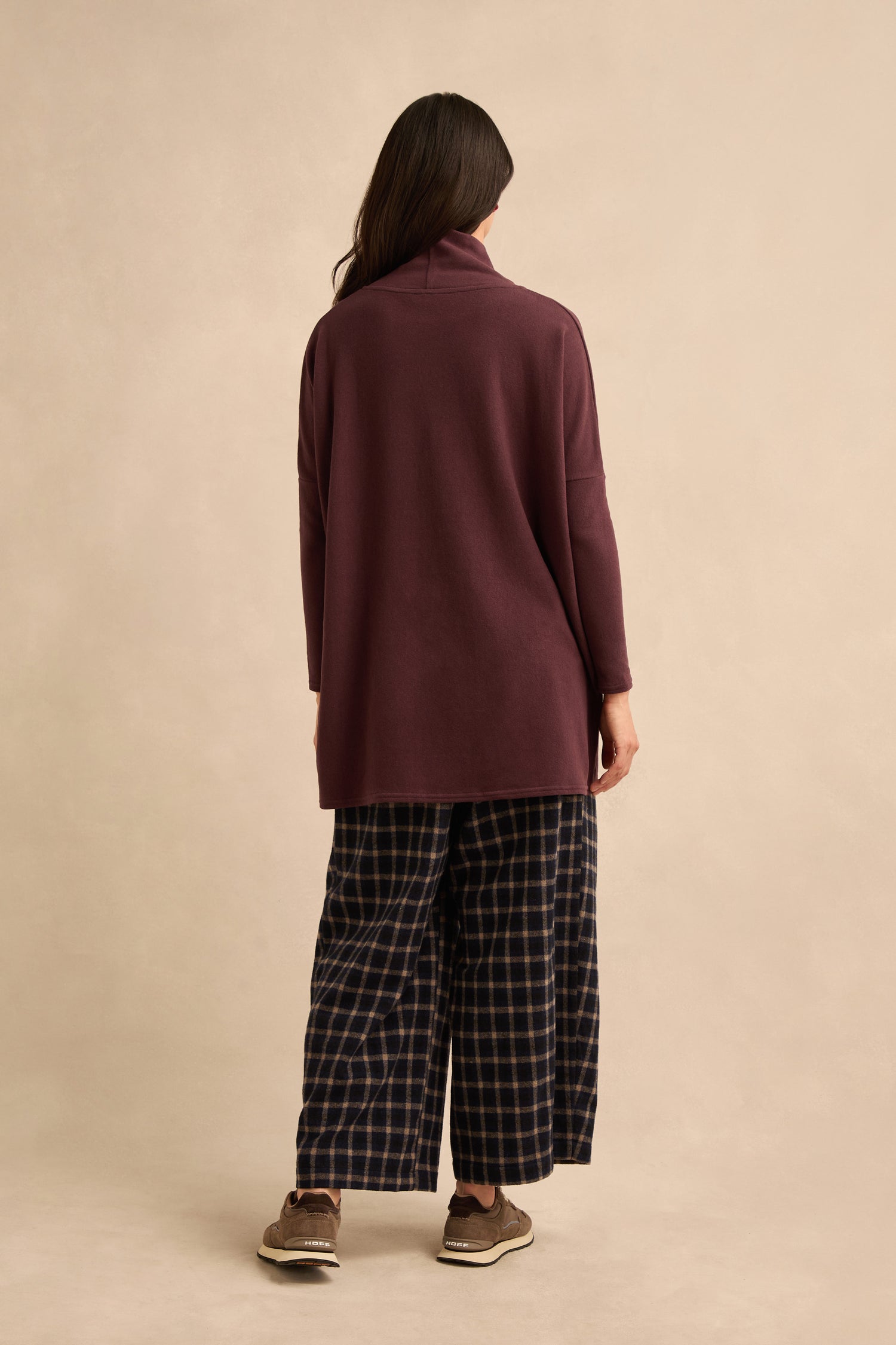 A person stands facing away, wearing the Cowl Neck Fig Fleece Tunic paired with dark plaid wide-leg pants and brown shoes, against a plain beige background.