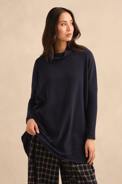 A person stands against a beige background wearing the Cowl Neck Navy Fleece Tunic, a loose-fitting style made in Italy, paired with plaid pants and looking slightly to the side.