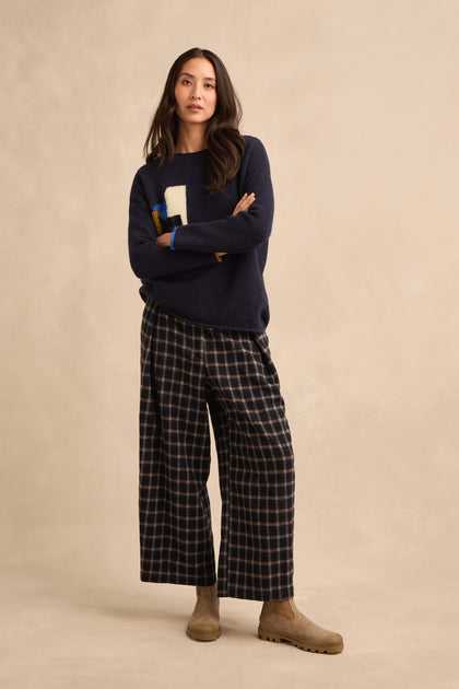 A woman stands with arms crossed, wearing the Patch Detail Jumper—a navy knit featuring a white “1” and abstract patch detail—paired with relaxed fit plaid wide-leg pants and beige suede boots against a neutral background.