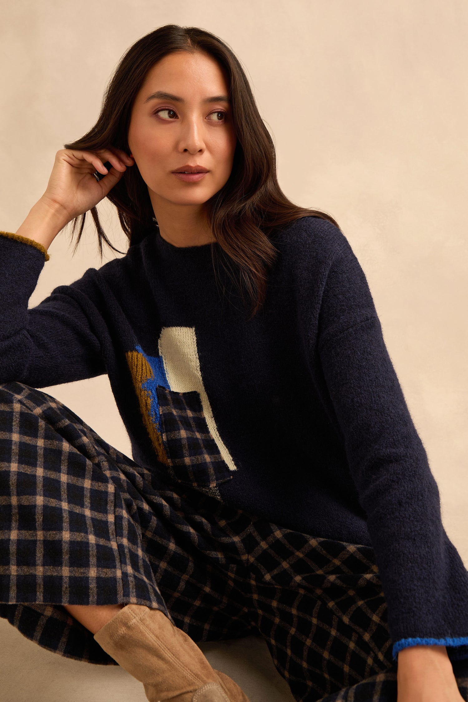A woman is seated, wearing the Patch Detail Jumper—a navy knit with an abstract patch design and relaxed fit—paired with plaid pants and beige boots, looking to the side against a neutral background.