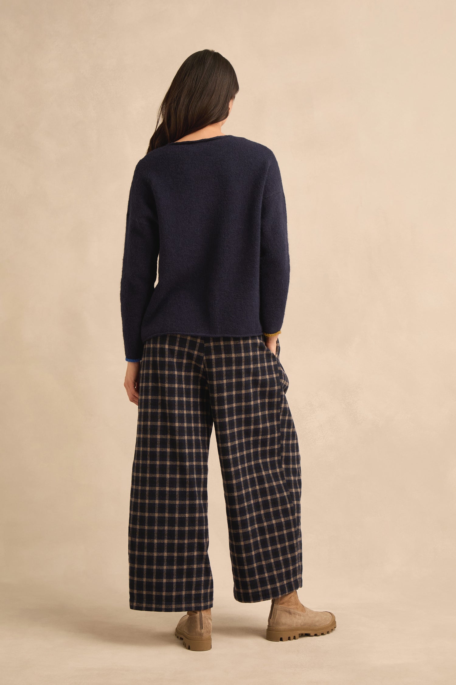 A person stands with their back to the camera, wearing the Patch Detail Jumper—a relaxed fit navy knit featuring an abstract patch—paired with wide-legged checkered pants and tan boots against a light beige background.