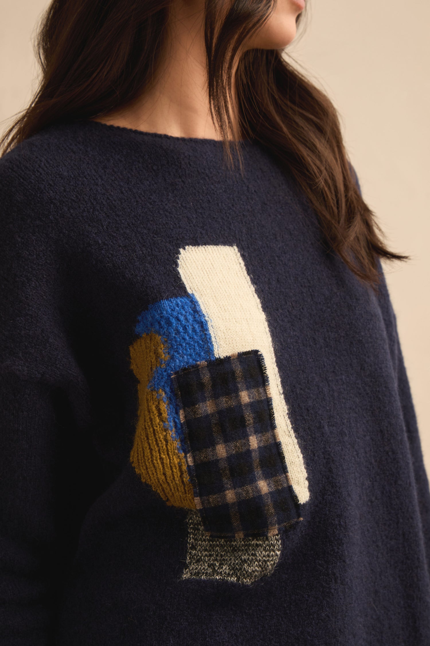 Wearing the Patch Detail Jumper, a relaxed fit navy knit featuring an abstract patchwork design with blue, yellow, white, and plaid fabric elements on the front.