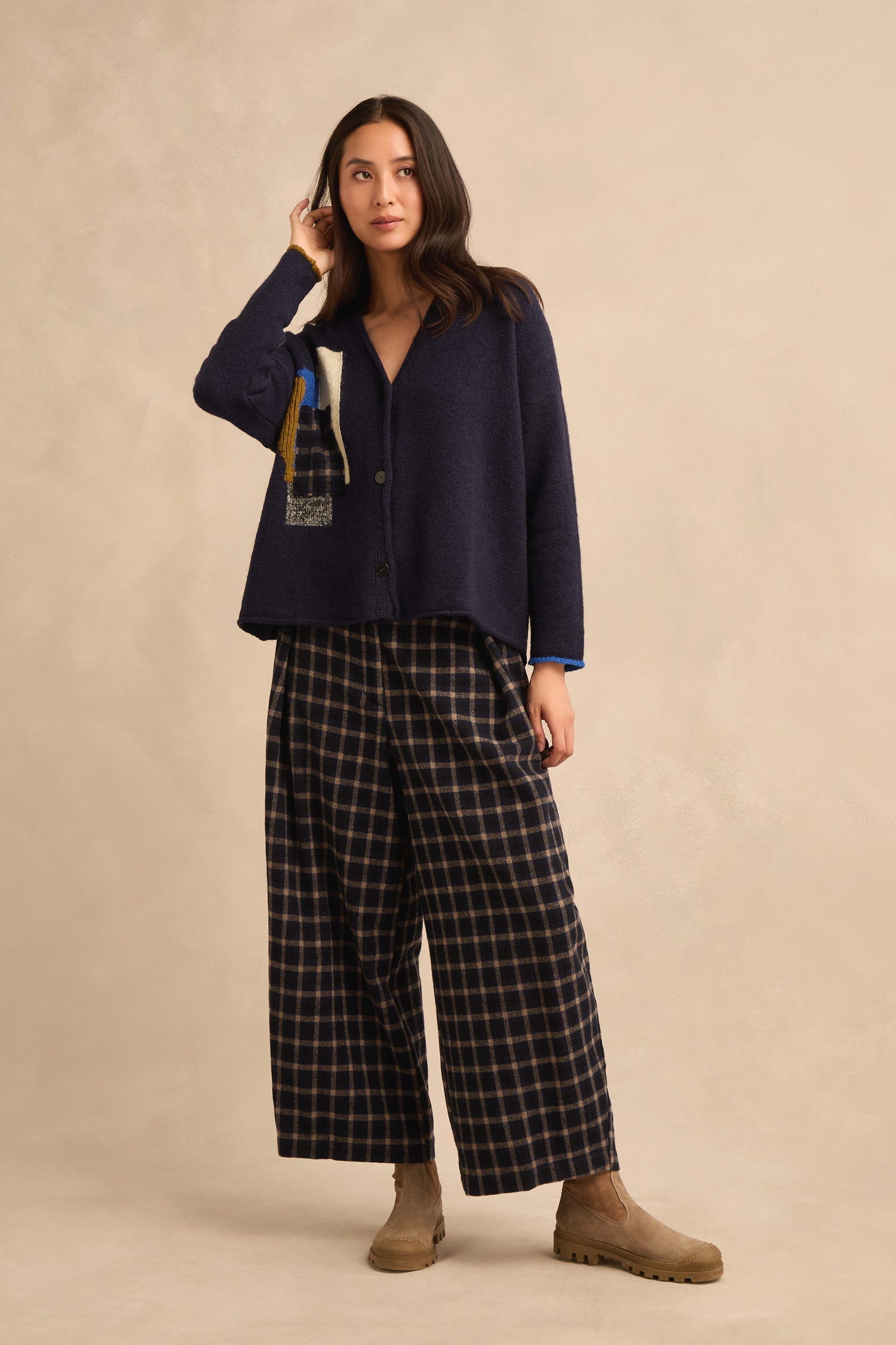 A woman wearing the Patch Detail Cardigan in navy, styled with wide-leg plaid pants and tan ankle boots, stands against a beige backdrop.
