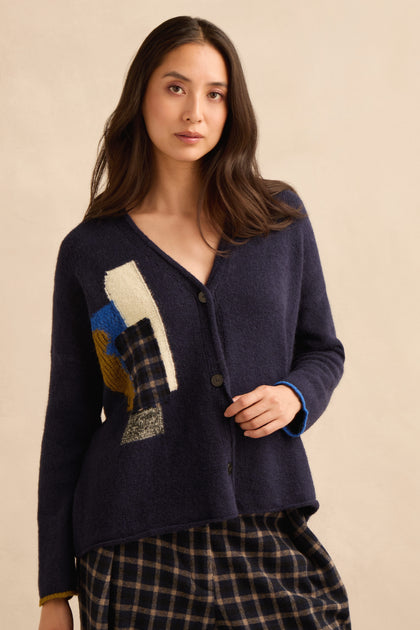 A woman stands against a plain beige background wearing the Patch Detail Cardigan—a navy knit cardigan with abstract patch details—paired with plaid pants. Made in Italy, this unique piece offers an elevated touch.