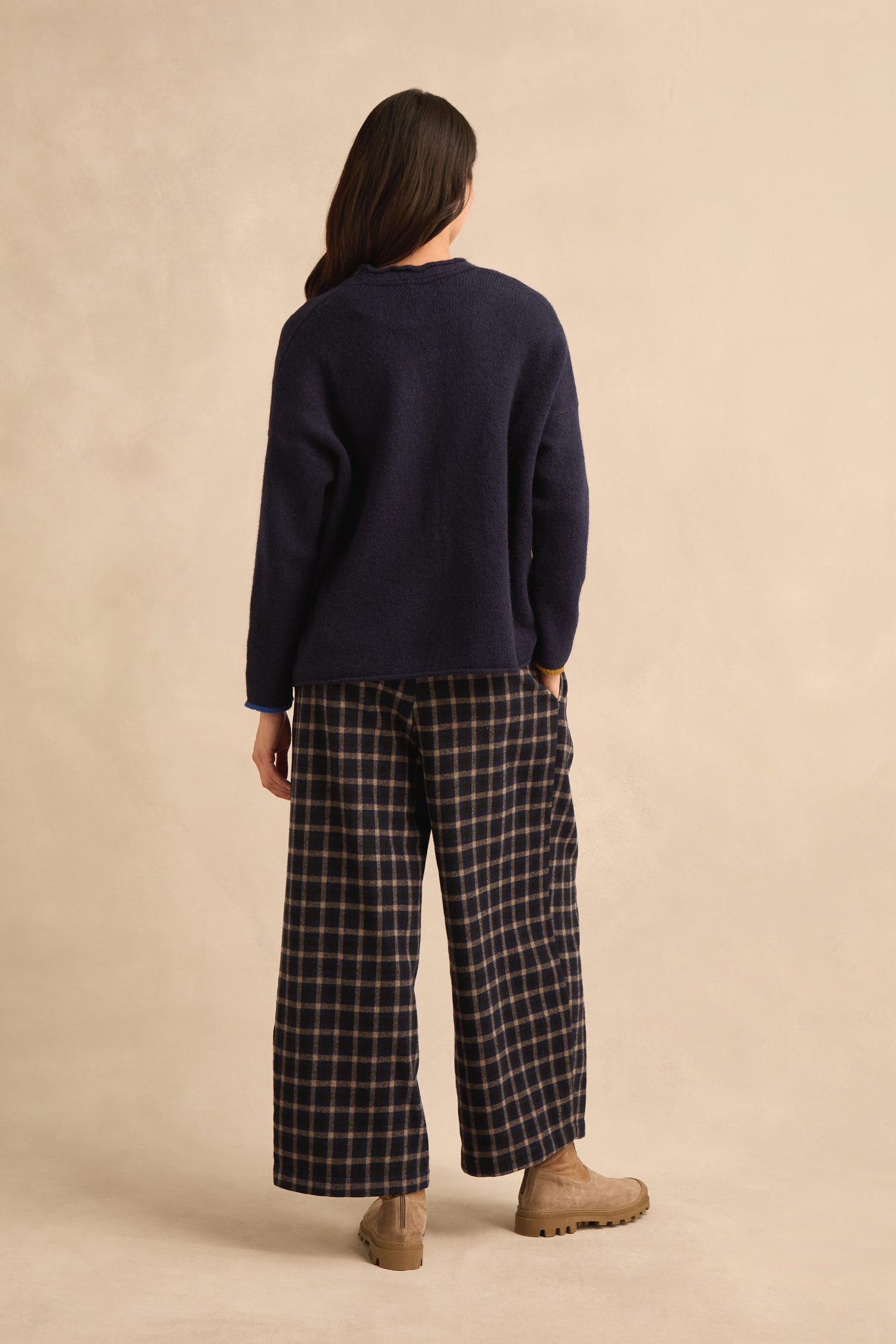 A person stands facing away against a neutral beige background, wearing the Patch Detail Cardigan in navy, paired with wide plaid pants and tan shoes.