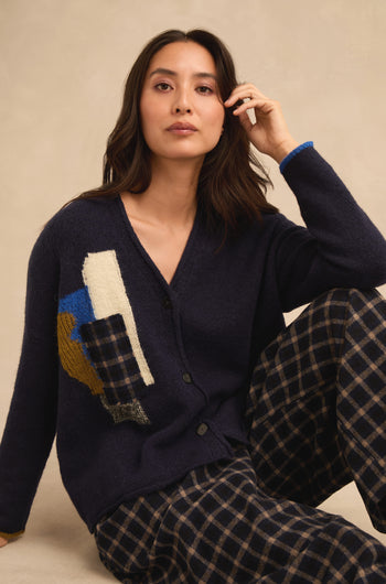 A woman is seated against a neutral background, wearing the Patch Detail Cardigan in navy with abstract patterns and plaid pants. Made in Italy, this stylish cardigan features patch details that showcase its sophisticated design and quality craftsmanship.