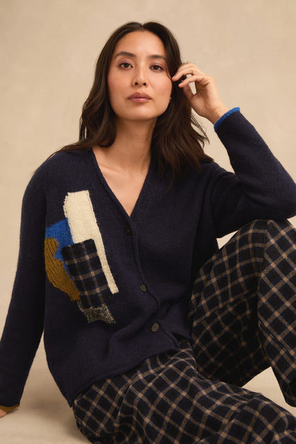 A woman is seated against a neutral background, wearing the Patch Detail Cardigan in navy with abstract patterns and plaid pants. Made in Italy, this stylish cardigan features patch details that showcase its sophisticated design and quality craftsmanship.