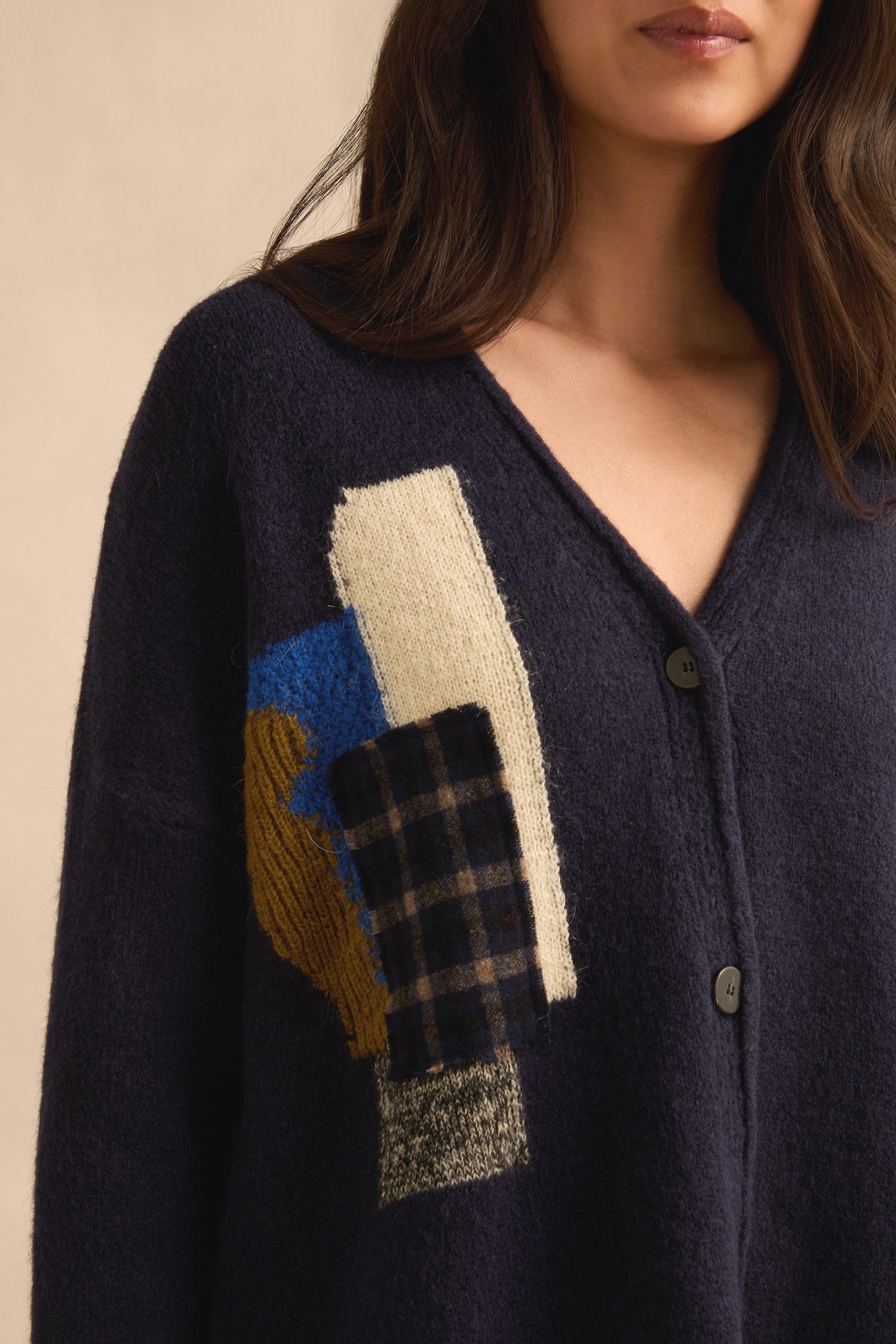 A woman is pictured wearing the Patch Detail Cardigan, a navy knit piece featuring abstract geometric patches in beige, blue, brown, and plaid on the left side. Made in Italy, only the lower half of her face and torso are visible.