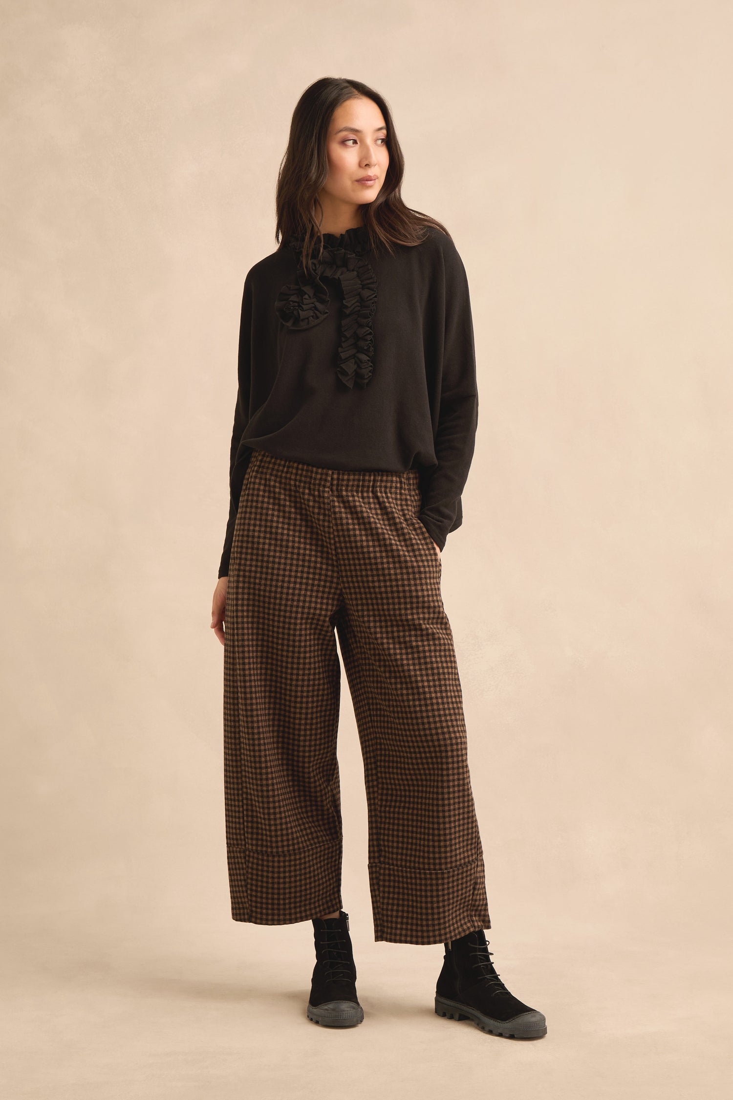 Woman standing against a beige background wearing a black long-sleeve top with ruffle details, Mini Check Jersey Bubble Trousers in a cropped length, and black boots.