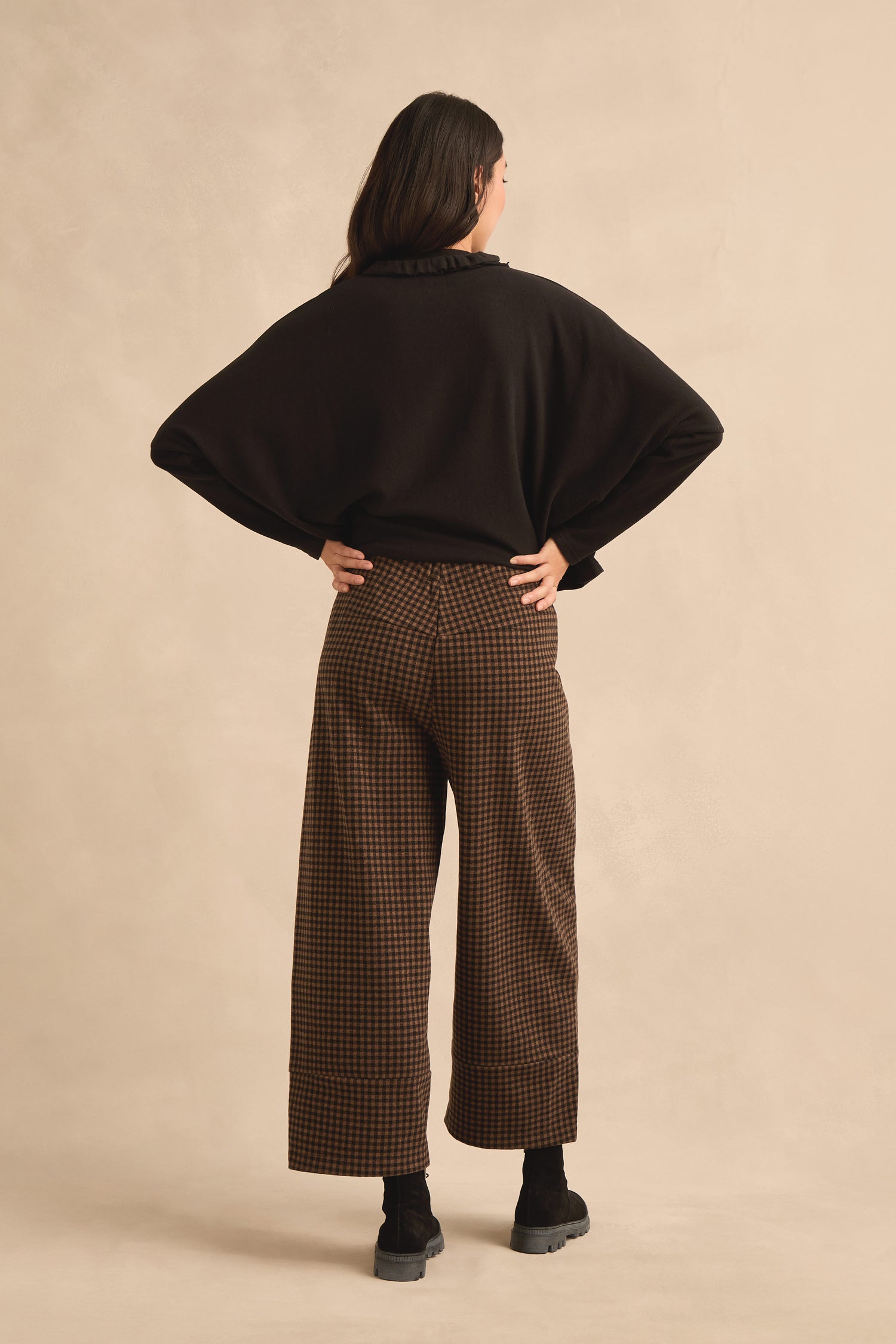 A person stands facing away, wearing a black long-sleeve top, black shoes, and the Mini Check Jersey Bubble Trousers against a beige background.