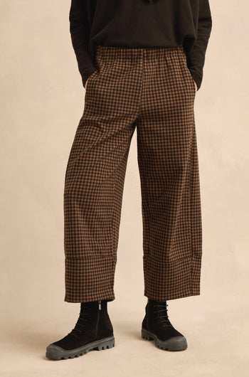 Wearing the Mini Check Jersey Bubble Trousers in brown and black, featuring a wide-leg, cropped silhouette, styled with black boots and a black top; the person stands with hands in pockets against a plain background.