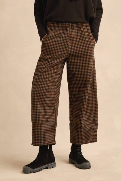 Wearing the Mini Check Jersey Bubble Trousers in brown and black, featuring a wide-leg, cropped silhouette, styled with black boots and a black top; the person stands with hands in pockets against a plain background.