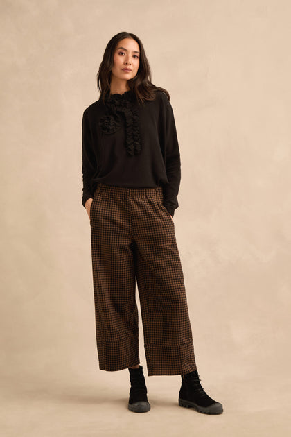 Woman standing against a beige backdrop, wearing a black ruffled sweater, Mini Check Jersey Bubble Trousers with a wide-leg fit in brown plaid, black socks, and black shoes, with hands in her pockets.