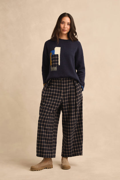 A woman stands against a beige background, wearing Tartan Pleated Trousers with a wide cropped leg in navy plaid, paired with a navy sweater featuring an abstract design and tan boots.