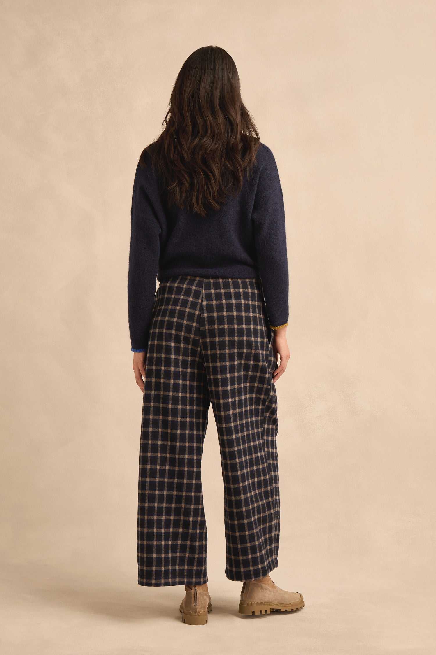 A person with long brown hair is shown from behind against a neutral background, wearing a navy sweater, tan boots, and the Tartan Pleated Trousers, which feature a wide cropped leg.