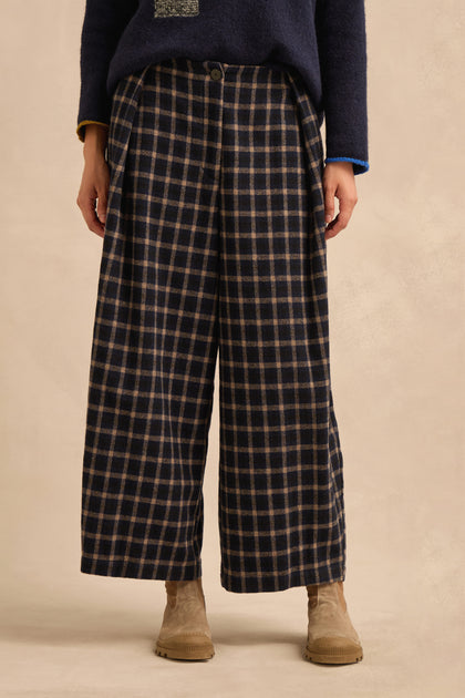 A person is shown from the waist down wearing Tartan Pleated Trousers featuring a wide cropped leg in navy and beige, paired with a navy sweater and tan suede boots. The background is plain and neutral.