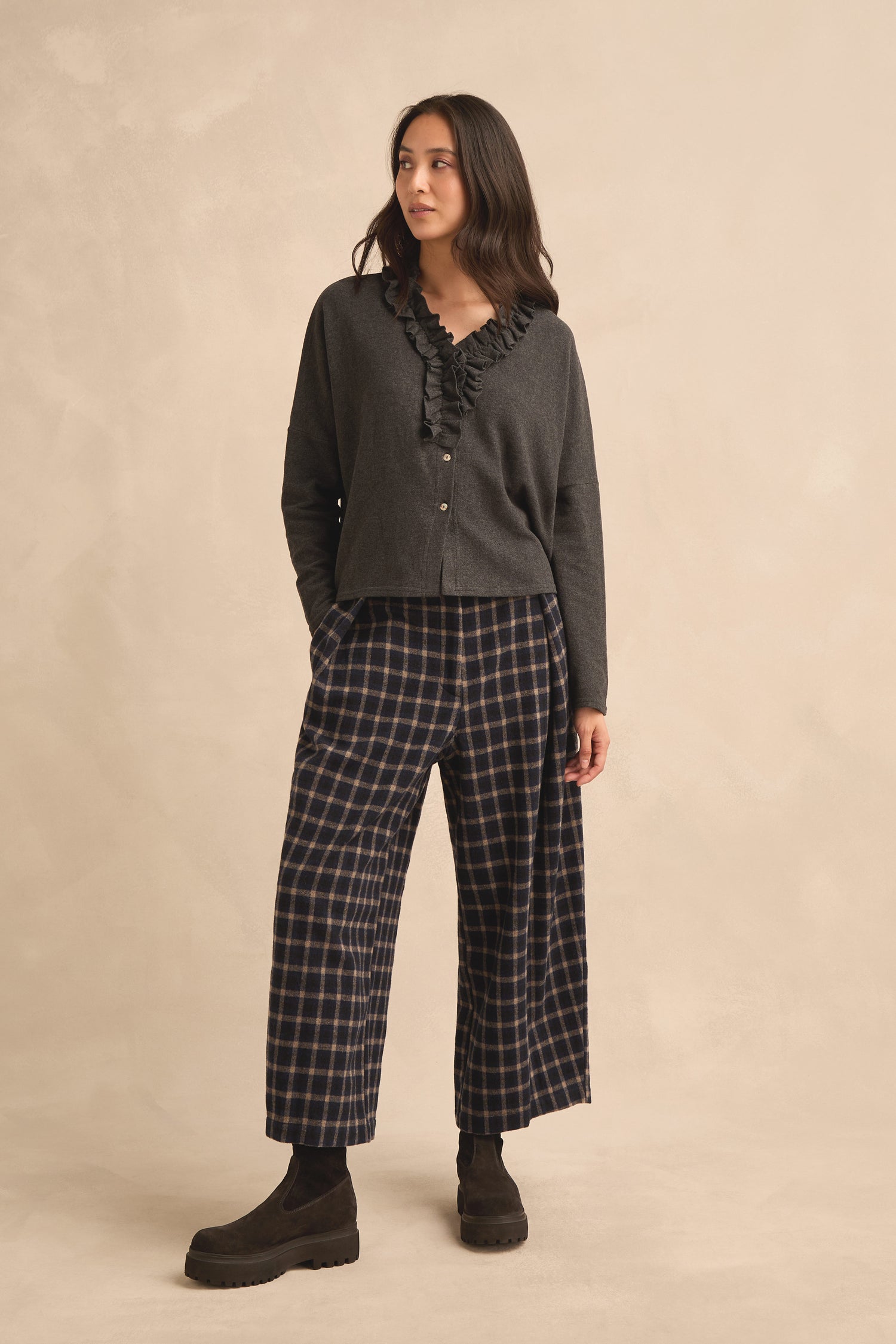 A woman stands against a light background with her hands in her pockets, wearing a Frill Neck Dark Grey Fleece Cardigan layered over a dark ruffled blouse, paired with plaid wide-leg pants and black boots.