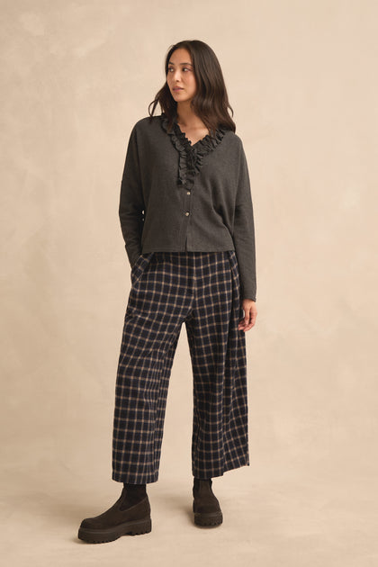 A woman stands against a light background with her hands in her pockets, wearing a Frill Neck Dark Grey Fleece Cardigan layered over a dark ruffled blouse, paired with plaid wide-leg pants and black boots.