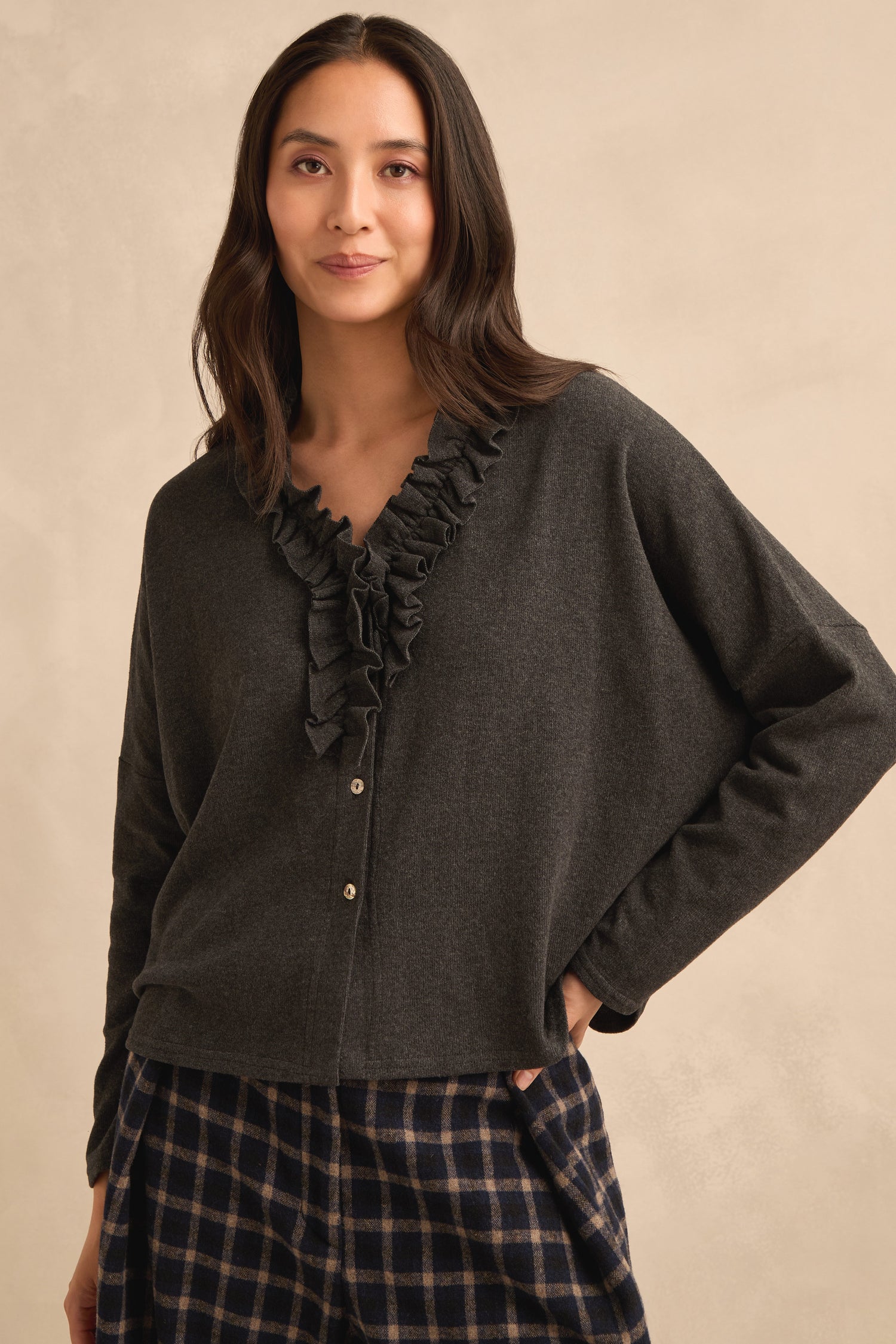 A woman wearing the Frill Neck Dark Grey Fleece Cardigan and plaid pants stands against a beige background with a relaxed expression.