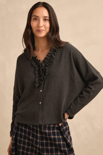 A woman wearing the Frill Neck Dark Grey Fleece Cardigan and plaid pants stands against a beige background with a relaxed expression.