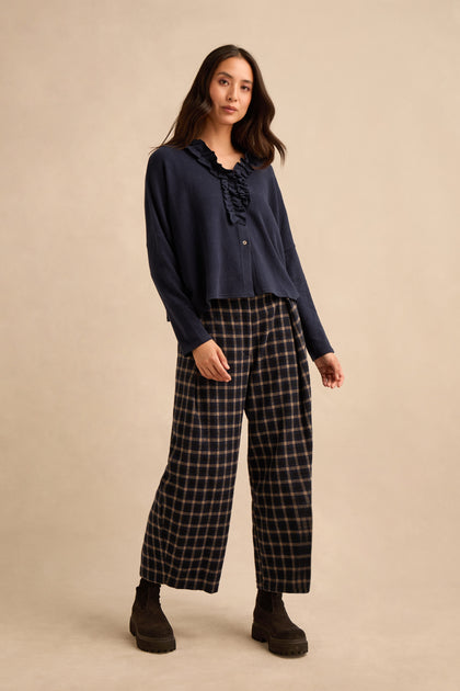 A woman stands against a plain beige background, wearing the Frill Neck Navy Fleece Cardigan layered over a ruffled blouse, wide-leg plaid pants, and dark boots.