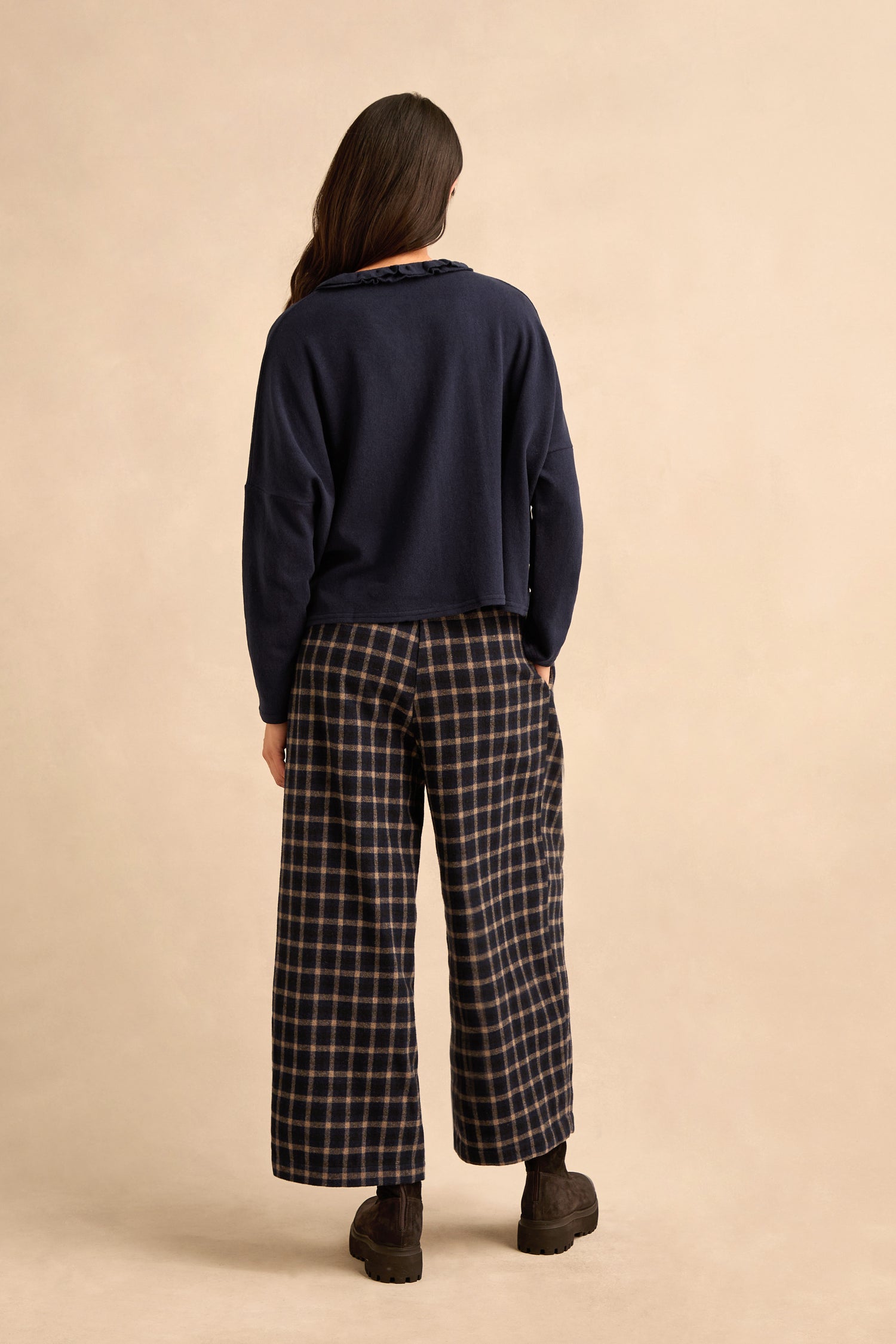 A person stands with their back to the camera, wearing the Frill Neck Navy Fleece Cardigan layered over a long-sleeve shirt, paired with wide-leg checkered pants and black boots against a plain beige background.