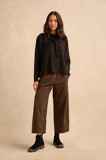 A woman stands against a beige backdrop, wearing the Black Frill Neck Top with ruffle details, brown plaid wide-leg pants, and black lace-up boots.