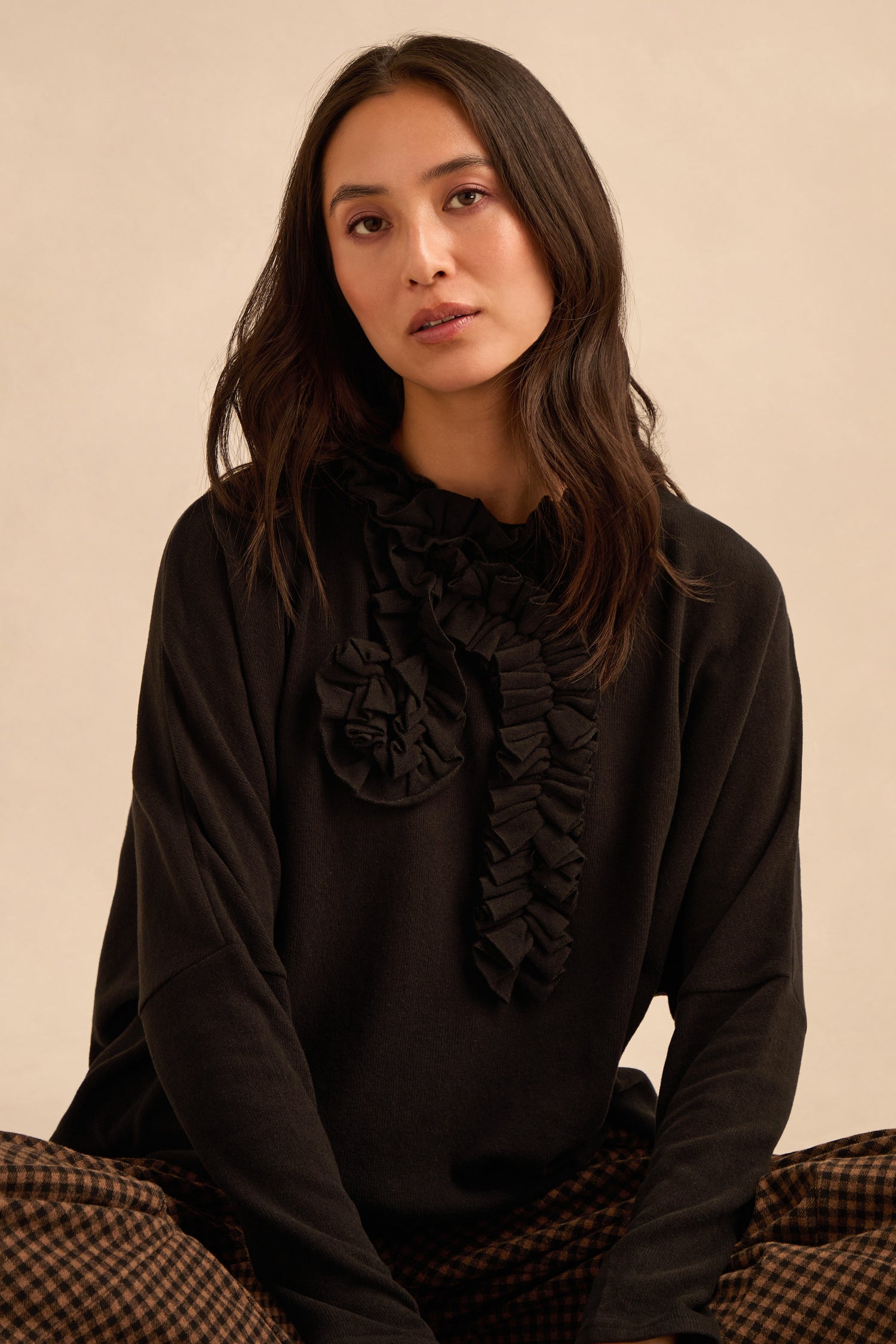 A woman with long dark hair wears the Black Frill Neck Top, made in Italy from black jersey with ruffled details, paired with checked pants as she sits against a neutral background.