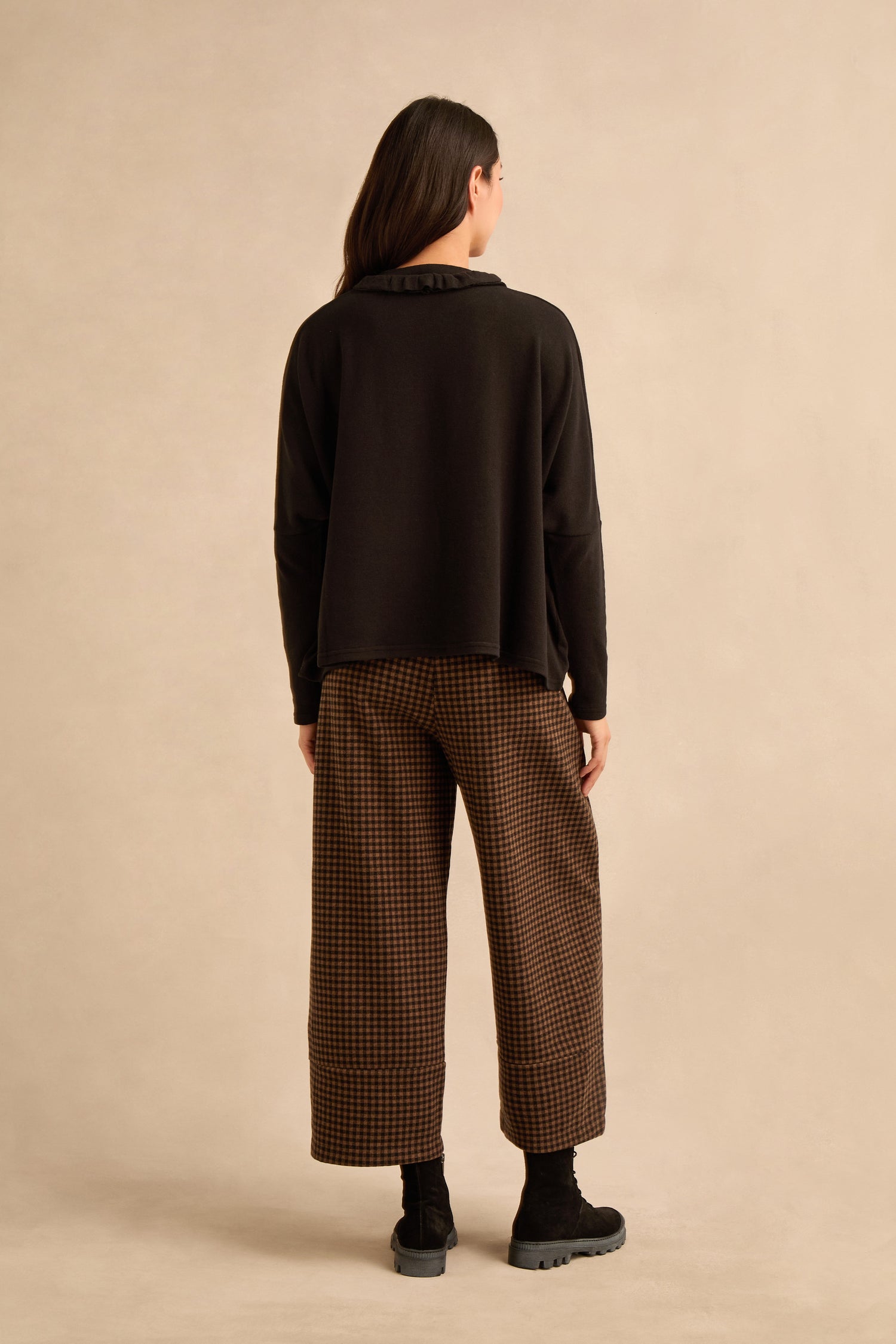 A person stands against a plain beige background, facing away, wearing the Black Frill Neck Top with long sleeves, brown checkered wide-leg pants, and black boots.