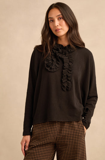 A woman with long dark hair and a neutral expression stands against a beige background, wearing the Black Frill Neck Top—crafted in Italy with a ruffled collar detail—and brown checkered pants.