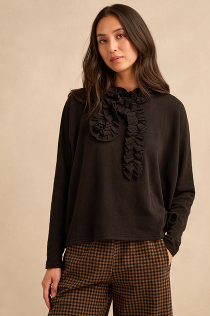 A woman with long dark hair and a neutral expression stands against a beige background, wearing the Black Frill Neck Top—crafted in Italy with a ruffled collar detail—and brown checkered pants.