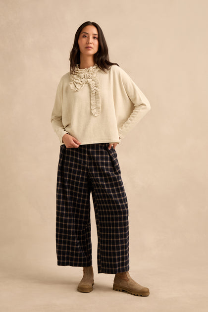A woman stands against a beige background wearing the Cream Frill Neck Top—a cream fleece jersey sweater with a relaxed, boxy fit and ruffle detail—paired with wide-leg plaid pants and tan boots.
