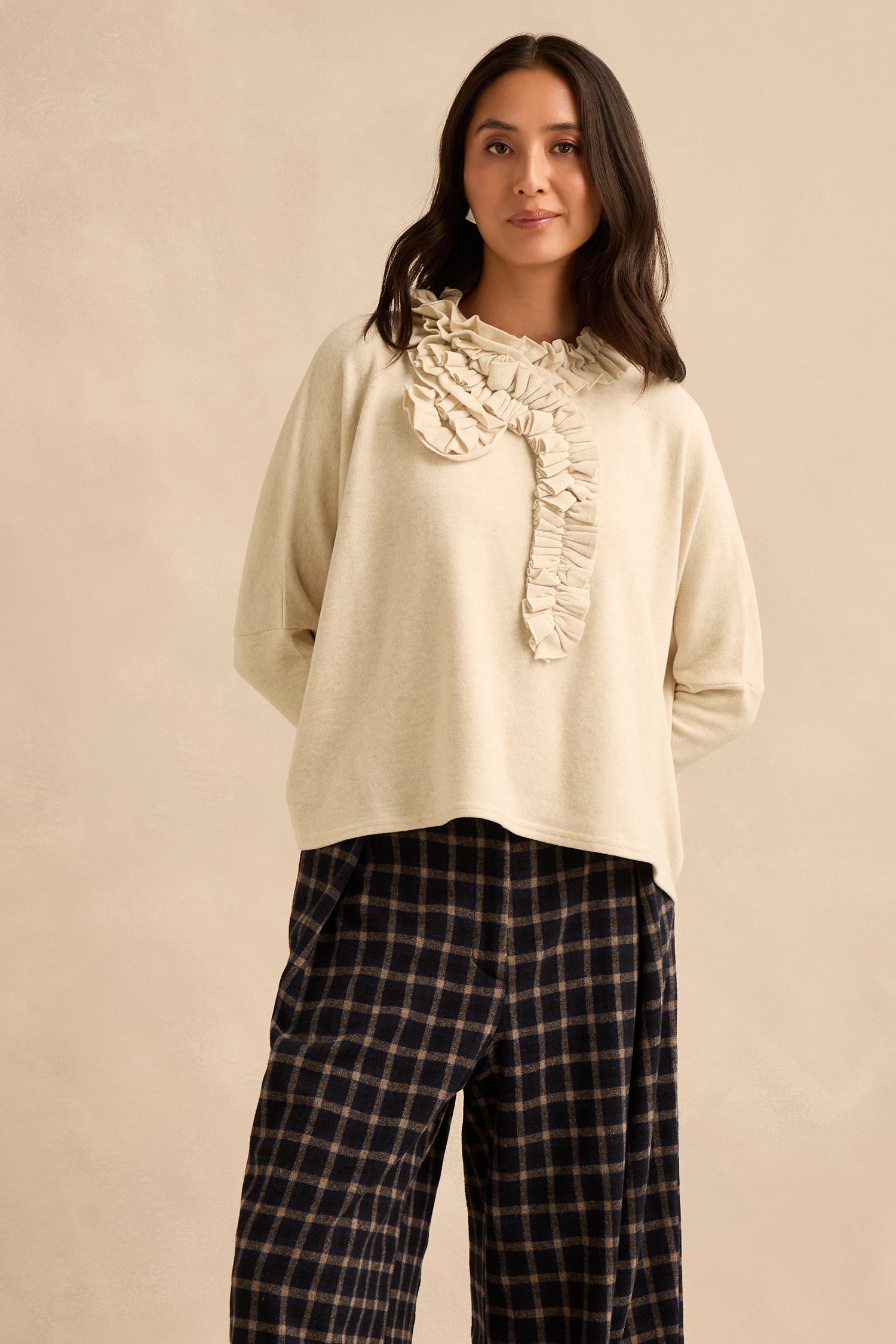 A woman stands against a neutral background, wearing the Cream Frill Neck Top with ruffle detail and black plaid pants. The top features a relaxed boxy fit, and she has her hands in her pockets.