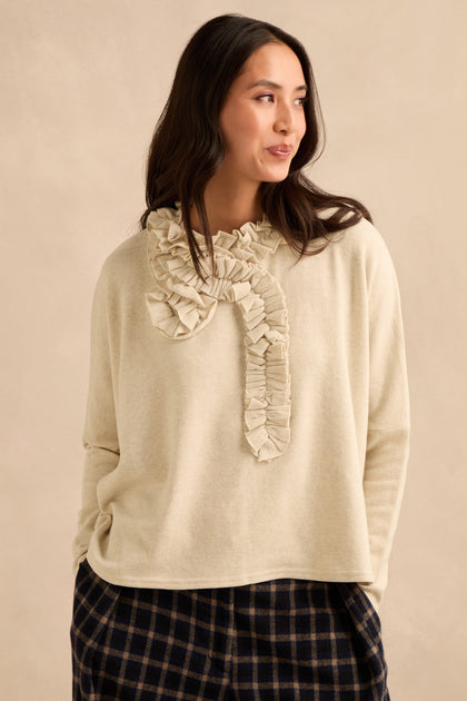 A woman is wearing the Cream Frill Neck Top, featuring a ruffled design at the neckline and a relaxed boxy fit, paired with dark plaid pants, as she stands against a neutral background.