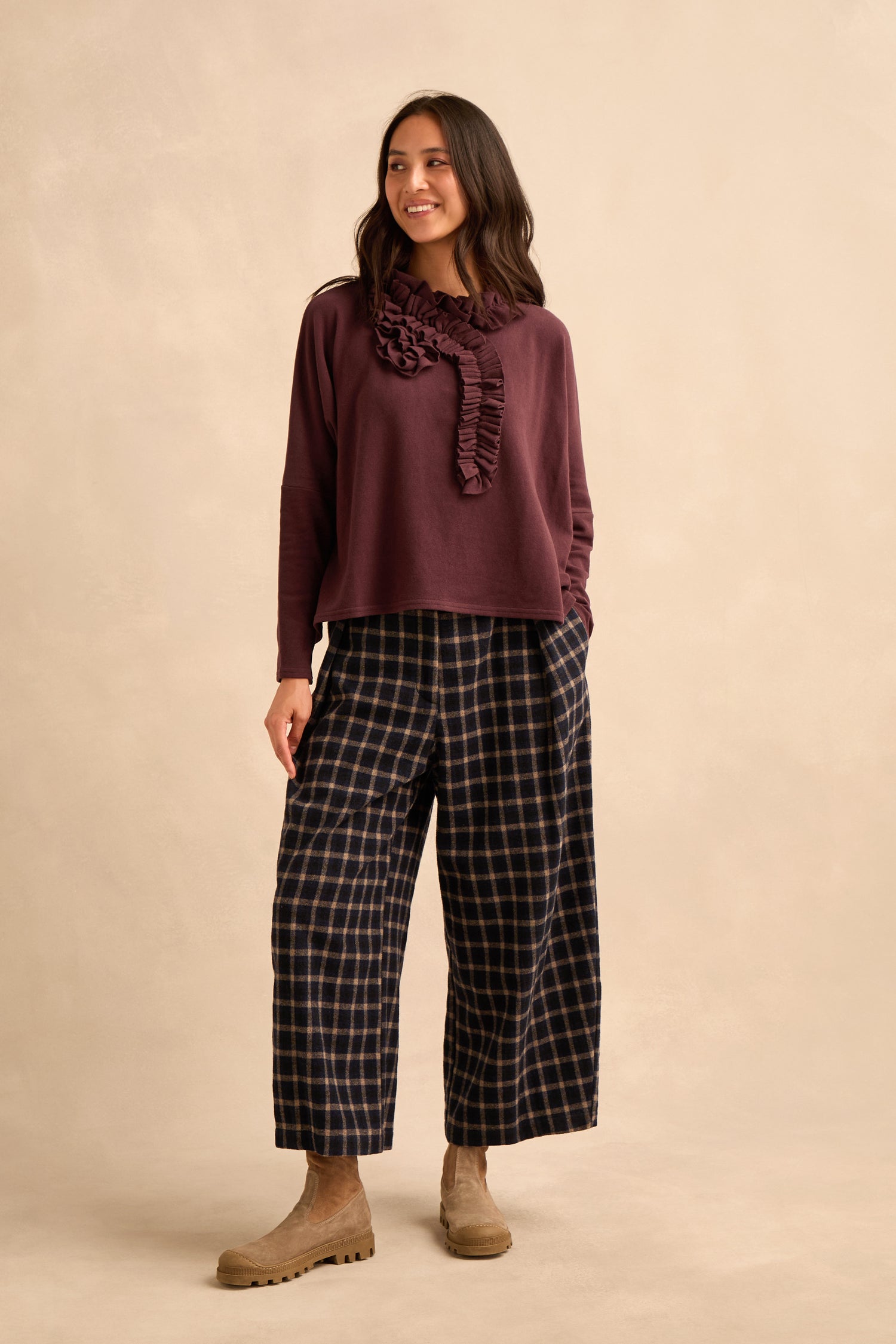 A woman stands against a beige background, wearing the Fig Frill Neck Top made in Italy, featuring ruffle details. She pairs it with wide-leg plaid pants and beige boots, her hands in her pockets and a slight smile on her face.