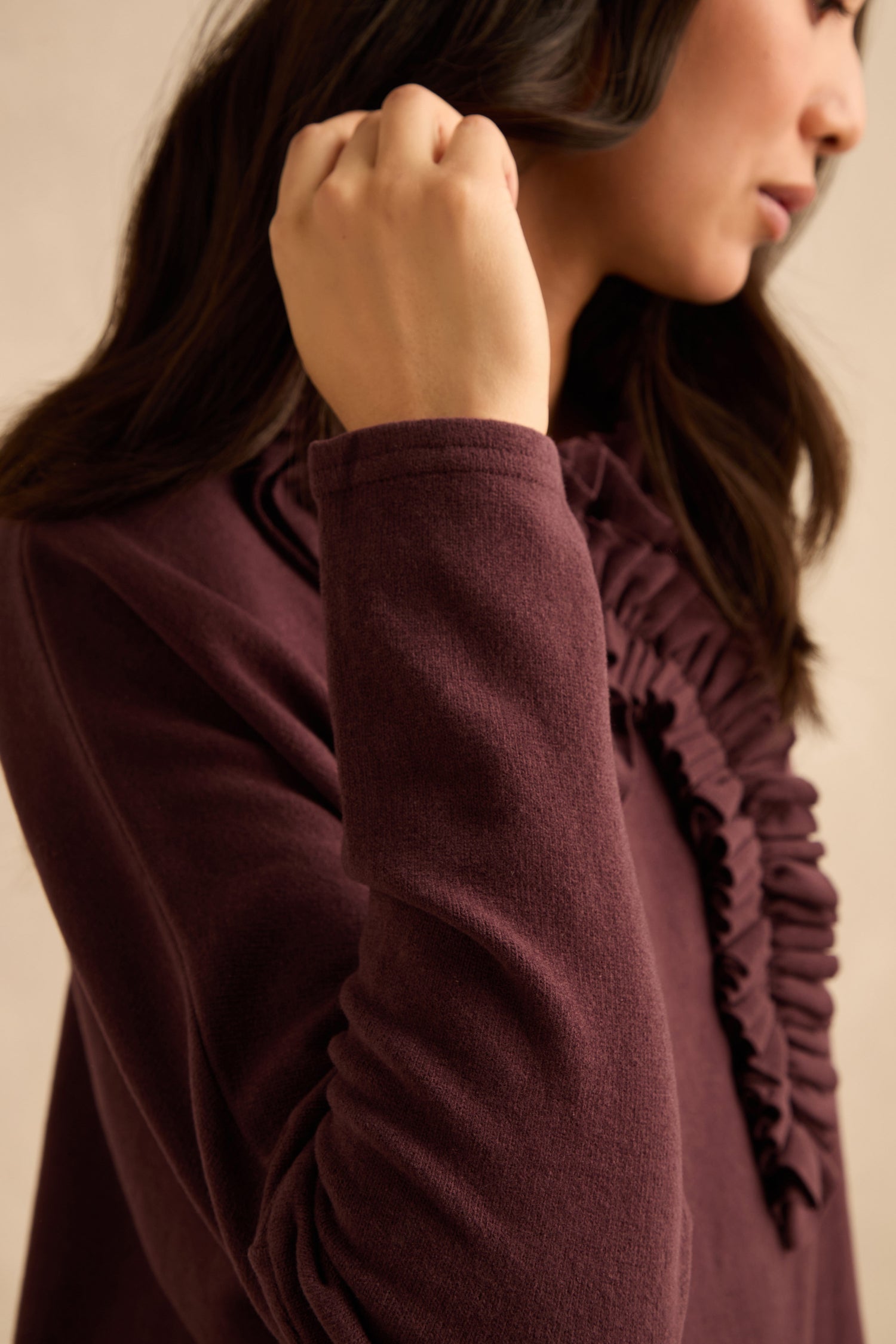A person with long dark hair wears the Fig Frill Neck Top, featuring ruffle details, raising their left hand near their face. The close-up side view photograph captures the area from shoulder to head.