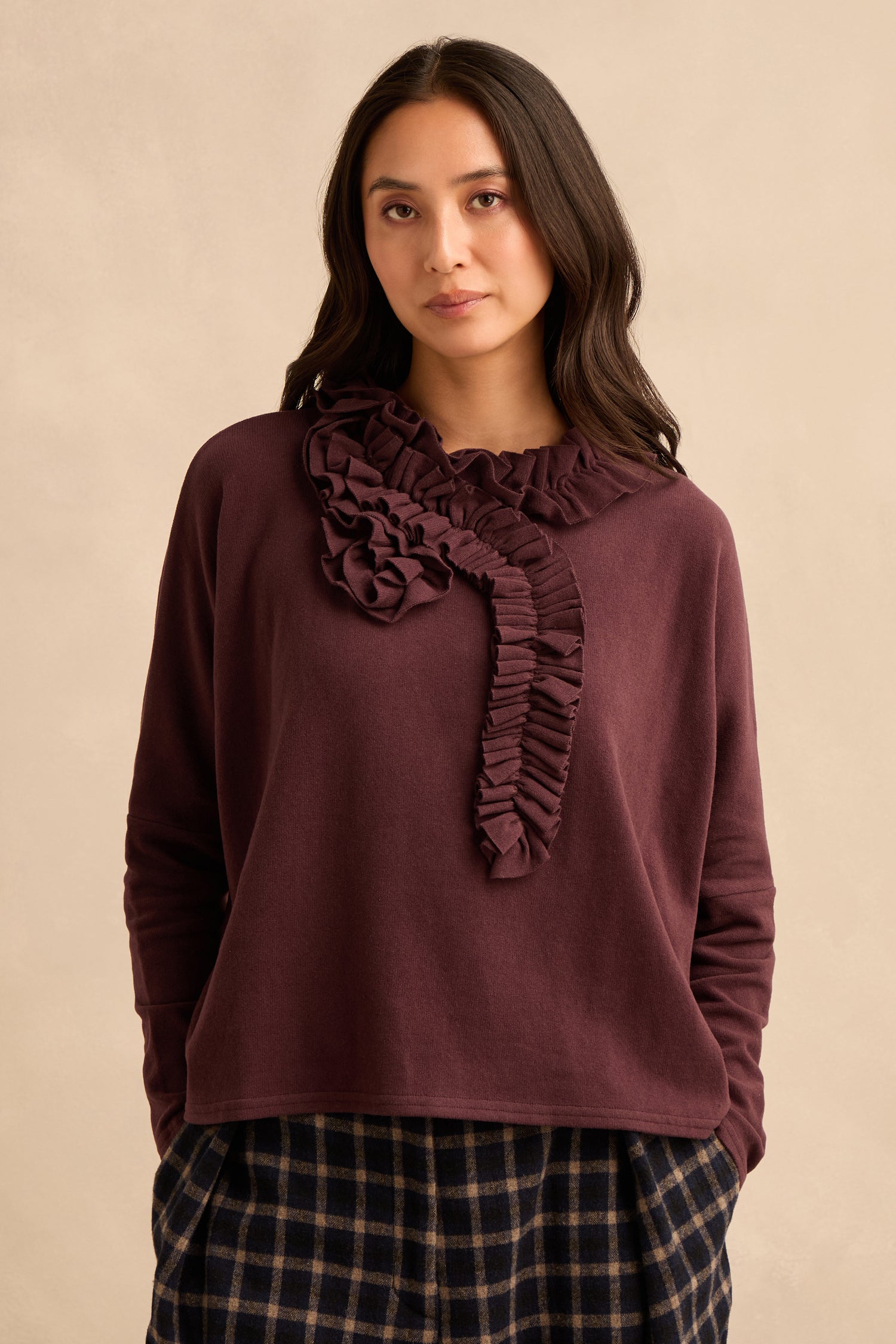A woman wears the Fig Frill Neck Top, a burgundy Italian-made piece featuring ruffled detailing at the neckline, paired with dark plaid pants and standing against a beige background.