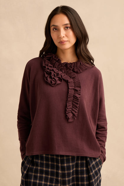A woman wears the Fig Frill Neck Top, a burgundy Italian-made piece featuring ruffled detailing at the neckline, paired with dark plaid pants and standing against a beige background.