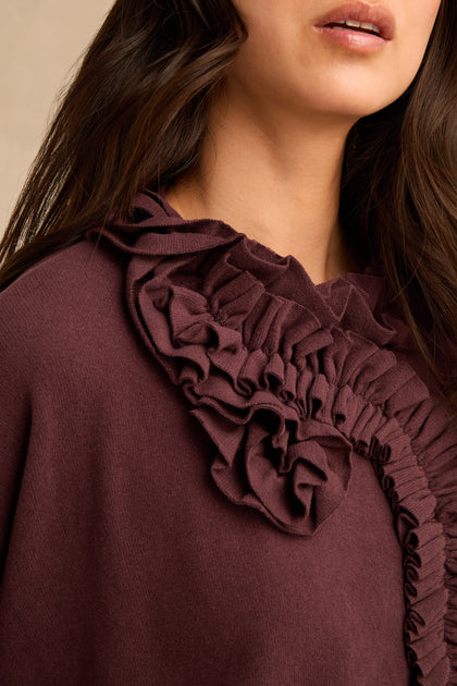 A woman shown from the shoulders up wearing the Fig Frill Neck Top, featuring a maroon hue and layered ruffle details near the neckline.