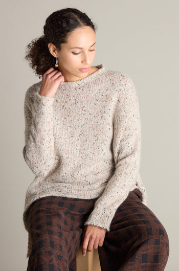 A person with curly hair is seated, wearing a Neirami knitted sweater and patterned pants, looking down with one hand touching the Marl Scoop Neck Knit.