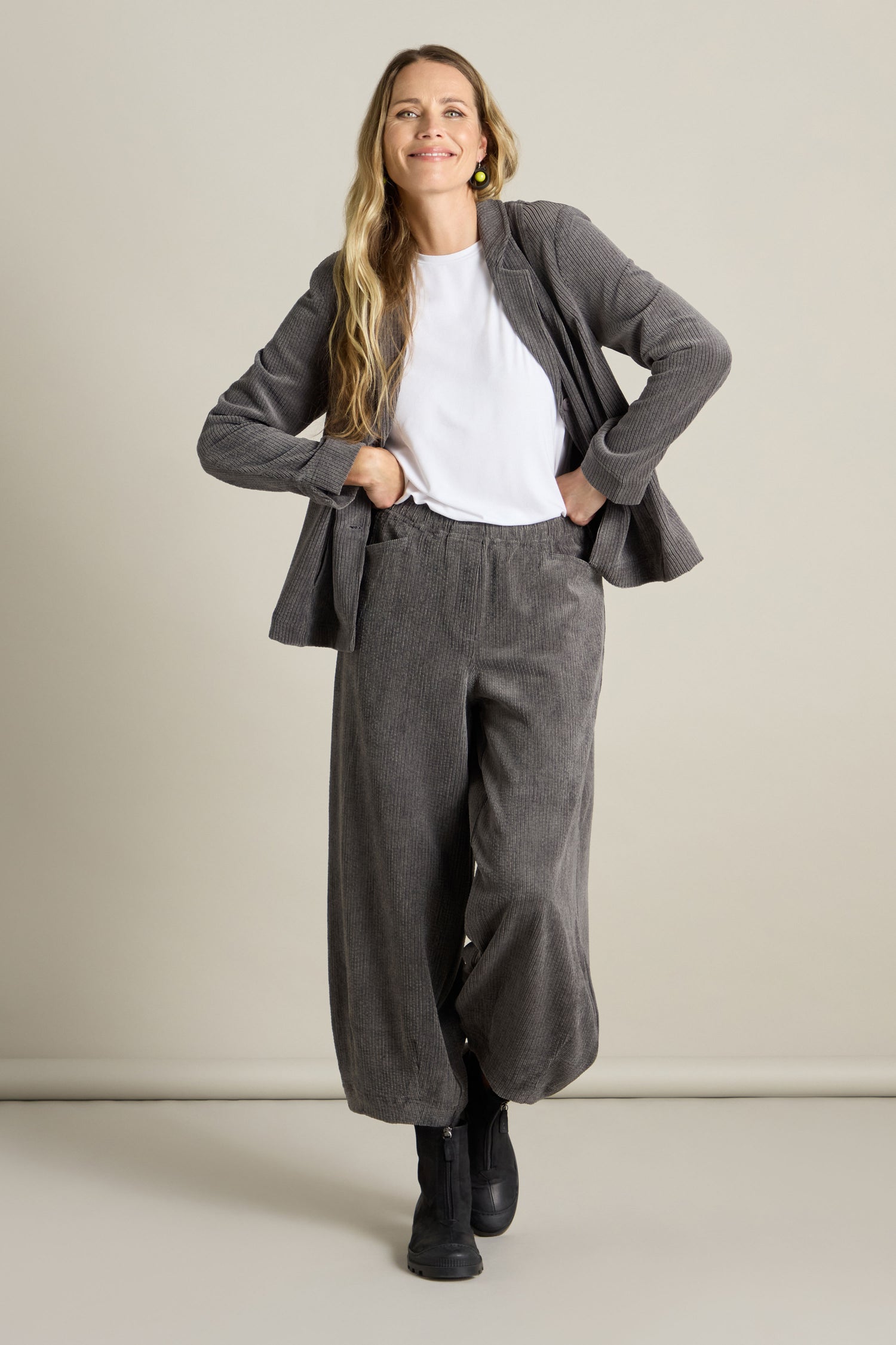 A woman wearing a white shirt and gray Stretch Cord Bubble Trouser, paired with a matching jacket and black shoes, stands against a plain background, smiling with her hands in her pockets.