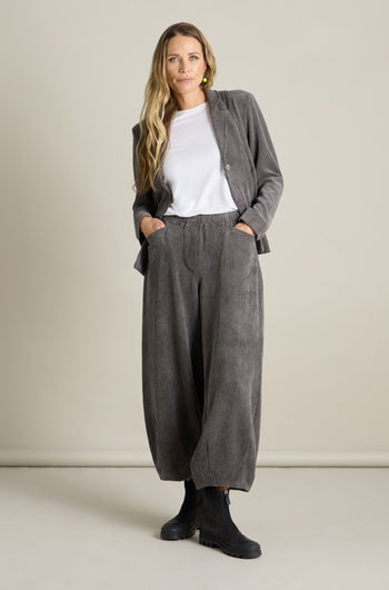 A person with long hair stands against a plain background, wearing the Stretch Cord Bubble Trouser paired with a white shirt and black boots. The trousers feature an elasticated waistband, adding comfort and style. They have one hand tucked in a pocket and are looking at the camera.