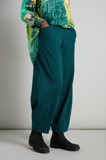 A person wears a patterned green top crafted from premium jersey fabric, paired with teal Jersey Bubble Trousers and black boots, set against a neutral backdrop with a touch of Danish-inspired style.